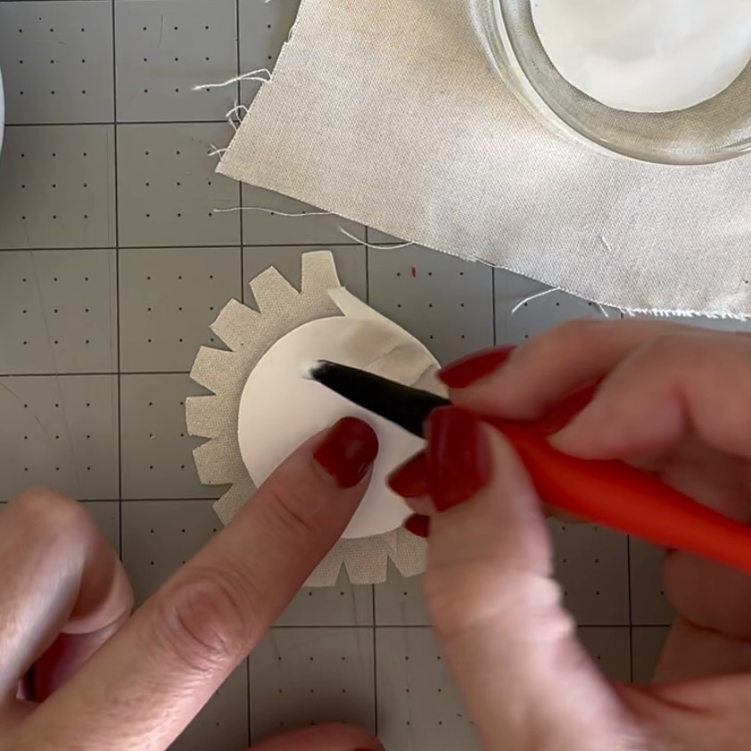 Applying glue to the back of a circular panel with clipped fabric tabs around the edge
